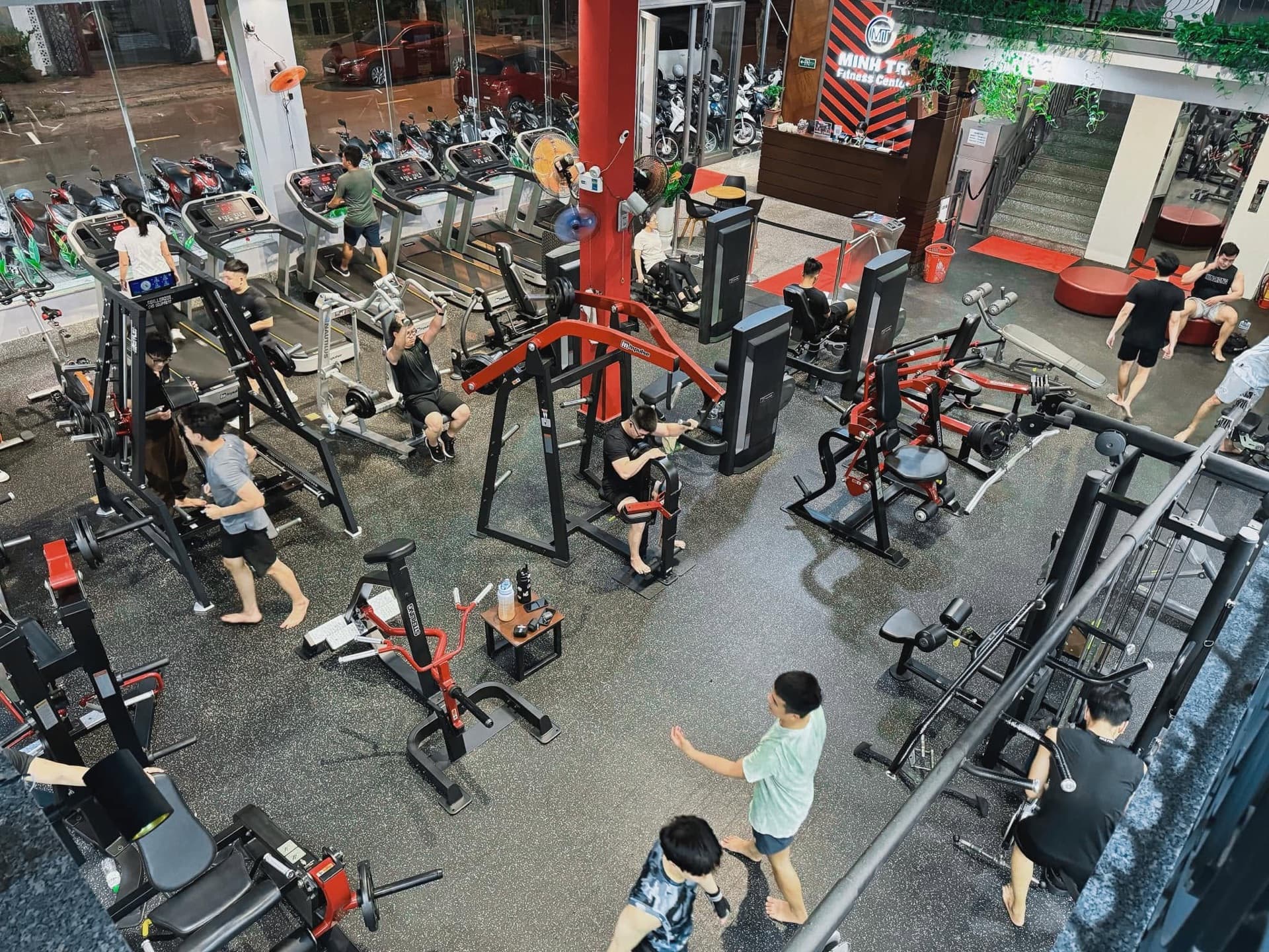 Sàn tập nhìn từ trên xuống buổi tối đông khách: dàn máy tạ đĩa ở giữa, quầy lễ tân với bảng hiệu Minh Trí phía cuối